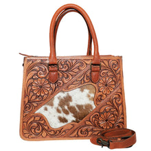 Load image into Gallery viewer, Brazos hand tooled leather tote, front view showing the hand-carved floral and scroll tooling framing a diagonal brown-and-white hair-on cowhide inset, with rolled leather handles and the removable crossbody strap alongside