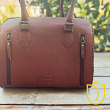 Load image into Gallery viewer, A brown pebbled leather bag with dual top handles, side zipper pockets, and a small embossed logo patch on the back, photographed outdoors on a wooden surface with blurred greenery in the background.