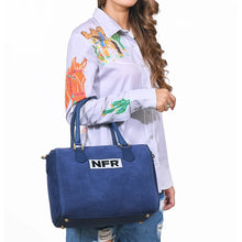 Load image into Gallery viewer, Lockhart navy suede Western speedy bag with NFR embroidered patch and rolled handles. A woman wearing a white embroidered long-sleeve shirt and ripped blue jeans carries a blue suede-like duffel bag with dark blue handles and a white patch reading 'NFR'.