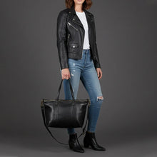Load image into Gallery viewer, Model in a black moto jacket carrying the Emmylou studded Italian leather tote in black by its handles, with the crossbody strap below
