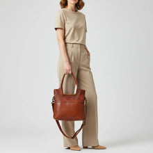 Load image into Gallery viewer, Model carrying the Sienna studded leather crossbody tote in cognac by its top handles, with the detachable strap hanging below
