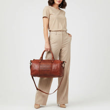 Load image into Gallery viewer, Model carrying the Rowan studded leather satchel in cognac by its top handles, with the detachable crossbody strap hanging below
