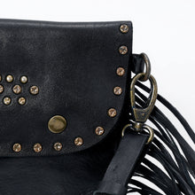 Load image into Gallery viewer, Close-up of the Estrella black leather crossbody flap showing antique-brass-tone studs, snap button, and swivel clasp beside the fringe
