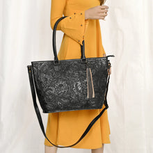 Load image into Gallery viewer, Model carrying the Maple Creek hand tooled leather tote in black on her forearm by its rolled handles
