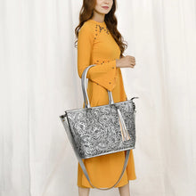 Load image into Gallery viewer, Model carrying the Maple Creek hand tooled leather tote in silver on her forearm by its rolled handles
