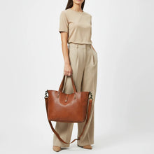 Load image into Gallery viewer, Model holding the Amber riveted leather crossbody tote in cognac by its handles, with the detachable strap hanging below

