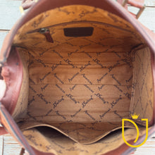 Load image into Gallery viewer, The interior of a brown leather handbag shown from above, featuring a tan patterned lining with repeating 'American Darling' text. A small dark brown label is visible near the top interior, and the bag is photographed against a light weathered wood background.
