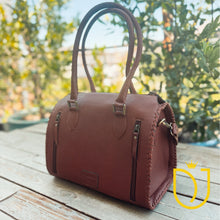 Load image into Gallery viewer, A brown leather handbag with double top handles and zipper compartments, placed on a wooden deck with green foliage in the background. A yellow watermark logo is visible in the bottom right corner.
