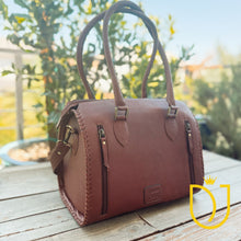 Load image into Gallery viewer, A brown textured leather handbag with double top handles and zippered pockets, resting on a wooden deck with a blurred garden background.
