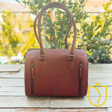 Load image into Gallery viewer, A brown leather handbag with dual top handles and zippered side pockets, displayed on a wooden surface with a blurred garden background.
