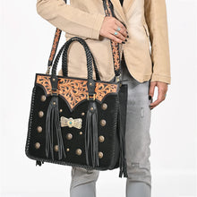 Load image into Gallery viewer, Fandango black hair-on leather tote held by its braided handles, showing the tooled yoke, brass conchos, turquoise-tone concho, and swinging fringe
