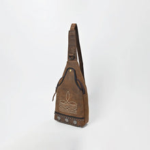 Load image into Gallery viewer, Austin western sling bag in distressed brown genuine leather, three-quarter view showing the tapered body, wide adjustable strap with D-ring, brass-tone snap at the top of the front panel, cream stitched sprig motif, and studded hem
